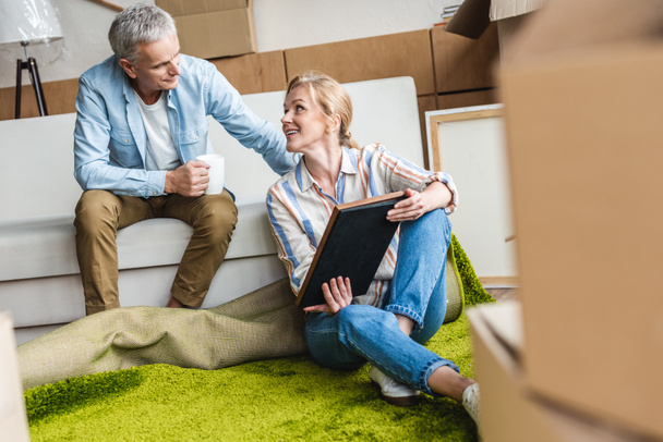 elderly couple looking at photo album while sitting between cardboard boxes in new house   - Photo, Image