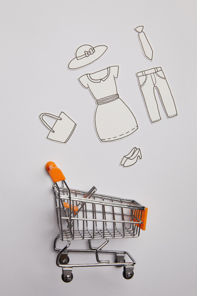 top view of shopping cart and little clothes made of paper on grey background - Foto, Imagen