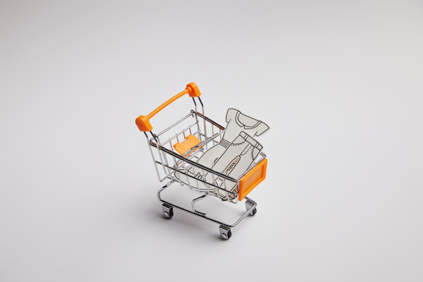 close up view of shopping cart with little clothes made of paper on grey background - Фото, изображение