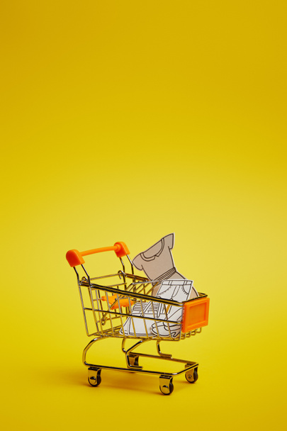 pohled z malého nákupního vozíku s papírové oblečení na žlutém pozadí na plochu - Fotografie, Obrázek