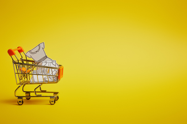 close up view of little shopping trolley with paper clothes on yellow backdrop - Photo, Image