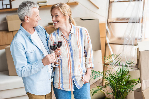happy senior couple holding glasses of wine and smiling each other during relocation - Photo, Image