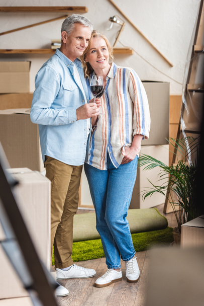 happy senior couple holding glasses of wine and looking away while moving home   - Photo, Image