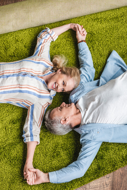 vista superior de feliz pareja de ancianos acostados juntos en la alfombra y tomados de la mano durante la reubicación
 - Foto, Imagen
