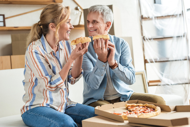 happy elderly couple eating pizza and smiling each other during relocation in new home - Photo, Image