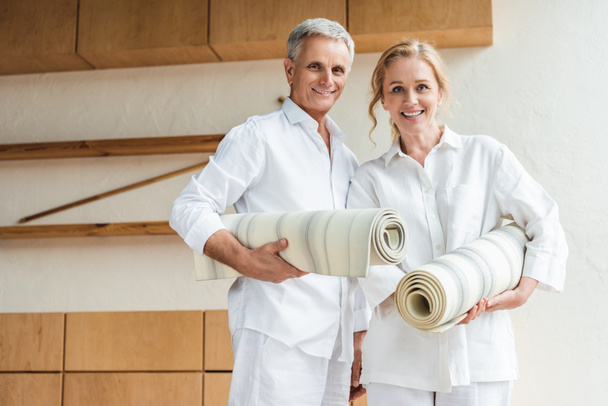 feliz casal sênior segurando tapetes de ioga e sorrindo para a câmera
 - Foto, Imagem