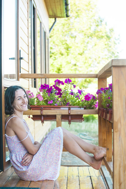 Das Mädchen sitzt auf dem Holzboden der Veranda. Eine barfüßige Frau ruht im Hof des Hauses. die Brünette im Sommerkleid sitzt vor der Haustür. - Foto, Bild