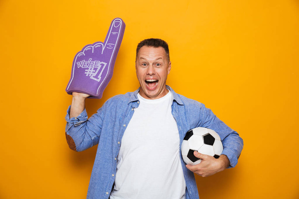 Image of excited man standing isolated over yellow background with football and fan glove. - 写真・画像