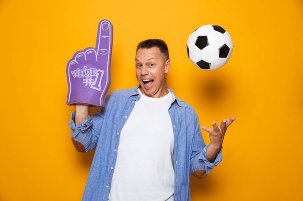 Image of excited man standing isolated over yellow background with football and fan glove. - Foto, Imagen