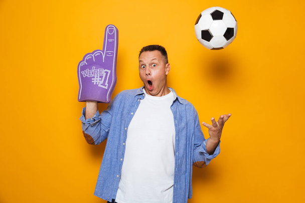 Image of shocked man with mouth opened standing isolated over yellow background with football and fan glove. - Foto, Imagem