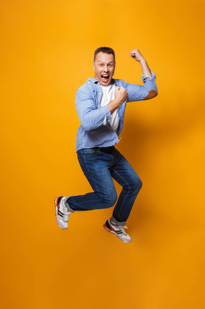 Image of emotional happy man jumping isolated over yellow background make winner gesture. - Φωτογραφία, εικόνα