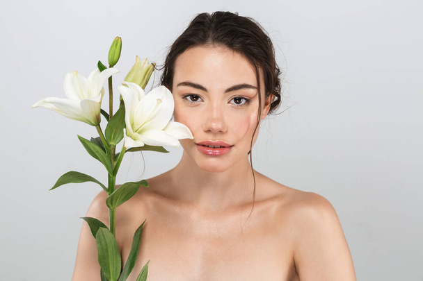 Beauty portrait of a lovely young topless woman with make-up posing with lily flower at her face isolated over gray background - 写真・画像