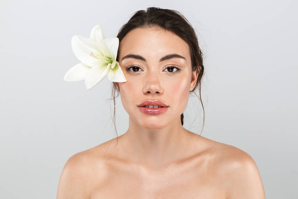 Beauty portrait of a pretty young topless woman with make-up posing with lily flower at her face isolated over gray background - Fotografie, Obrázek