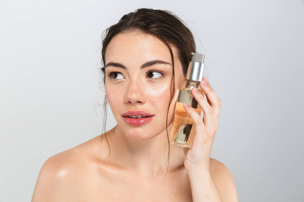 Beauty portrait of a cheerful young topless woman with make-up holding an oil bottle isolated over gray background - Fotografie, Obrázek