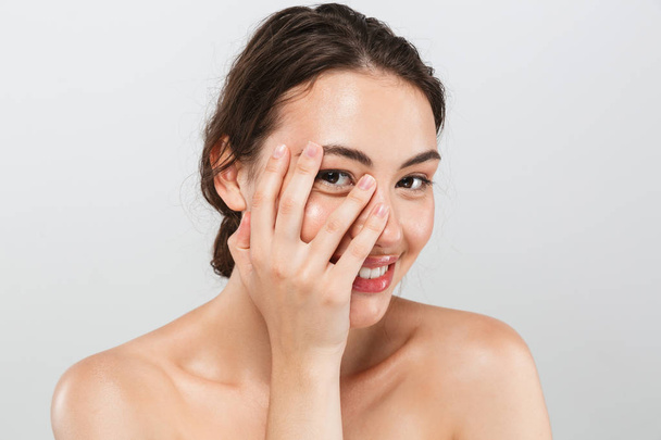 Retrato de belleza de una joven en topless sonriente con maquillaje mirando a la cámara a través de dedos aislados sobre fondo gris
 - Foto, Imagen