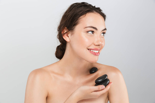 Beauty portrait of a lovely young topless woman holding spa hot massage stones isolated over gray background - 写真・画像