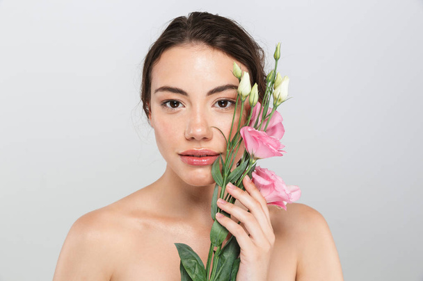 Beauty portrait of a lovely young topless woman with make-up posing with eustoma flowers at her face isolated over gray background - Foto, Bild
