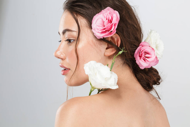 Side view beauty portrait of a pretty young topless woman with make-up posing with eustoma flowers at her face isolated over gray background - 写真・画像