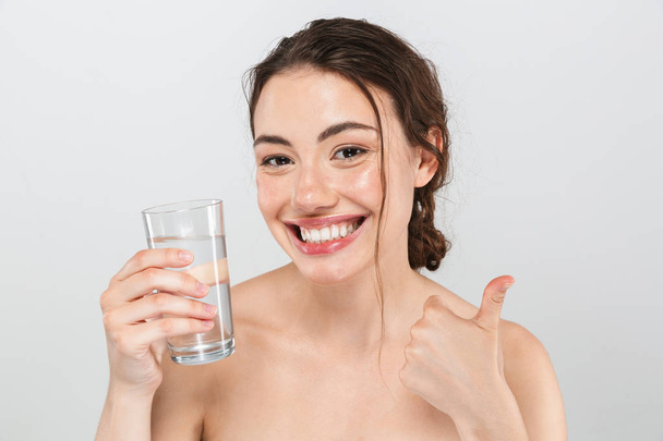 Beauty portrait of a smiling young topless woman with make-up holding glass of water and showing thumbs up isolated over gray background - Valokuva, kuva