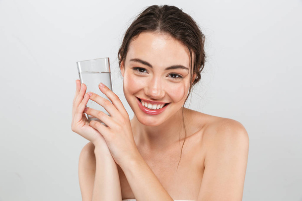 Beauty portrait of a happy young topless woman with make-up holding glass of water isolated over gray background - Фото, зображення