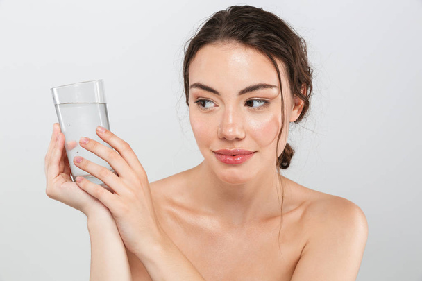Beauty portrait of a lovely young topless woman with make-up holding glass of water isolated over gray background - Photo, Image