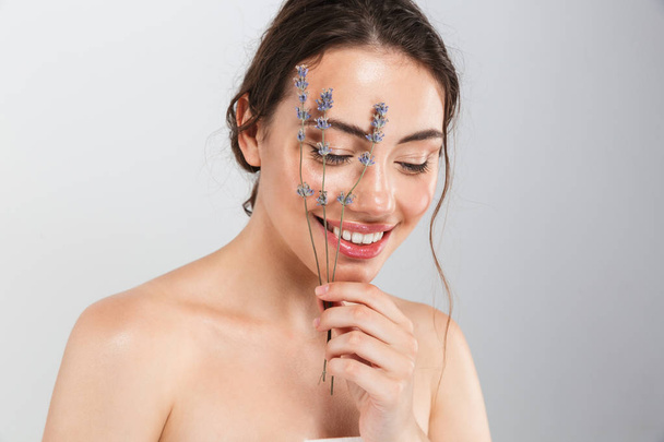 Beauty portrait of a smiling young topless woman with make-up posing with lavender flower at her face isolated over gray background - Foto, Bild