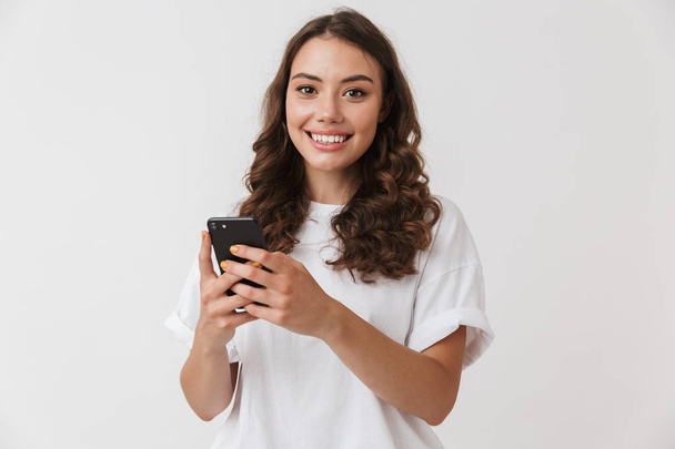 白い背景に分離された携帯電話を保持している笑顔の若いカジュアルなブルネット女性の肖像画 - 写真・画像