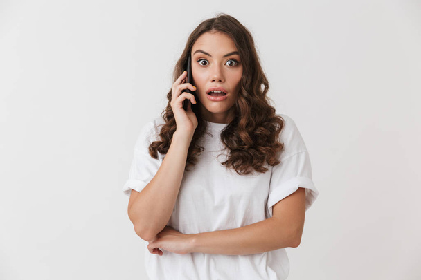 Portrait of a shocked young casual brunette woman talking on mobile phone isolated over white background - 写真・画像