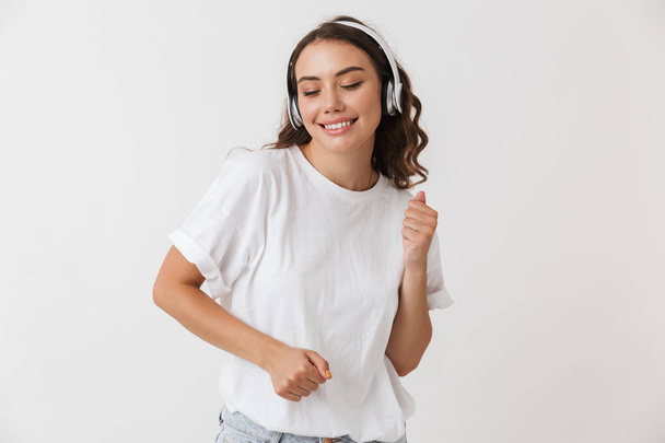 Portrait of a smiling young casual brunette woman listening to music with headphones isolated over white background - Photo, Image