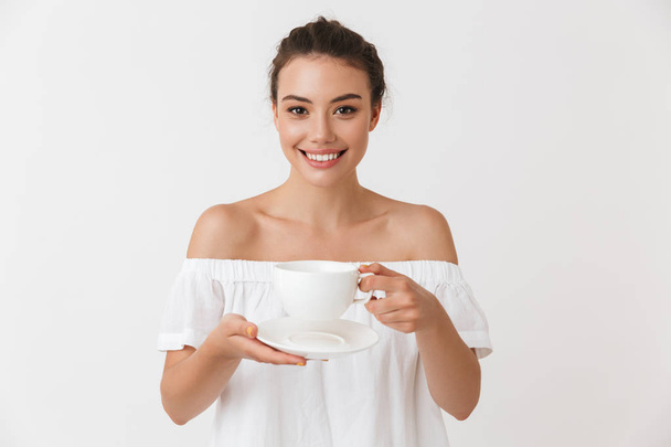 Portrait of a smiling young casual brunette woman holding cup of tea with saucer isolated over white background - Foto, imagen