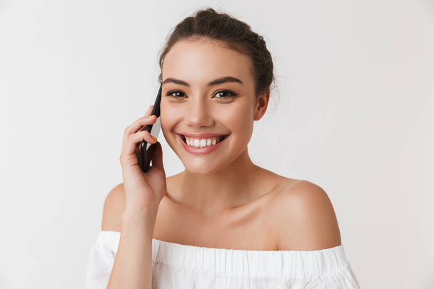 Portrait of a smiling young casual brunette woman talking on mobile phone isolated over white background - Фото, изображение