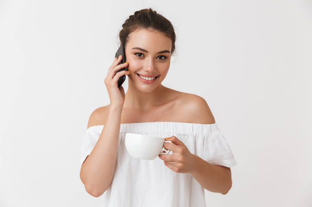Portrait of a smiling young casual brunette woman talking on mobile phone holding cup of coffee isolated over white background - Photo, image