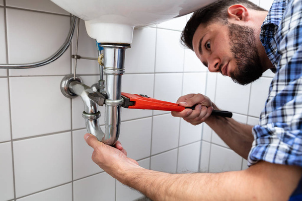Young Male Plumber Repairing Sink With Adjustable Wrench In Bathroom - Photo, Image