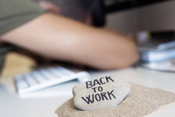 closeup of a concerned man sitting at his office desk, and stone on a pile of sand in the foreground with the text back to work written in it - Foto, Bild