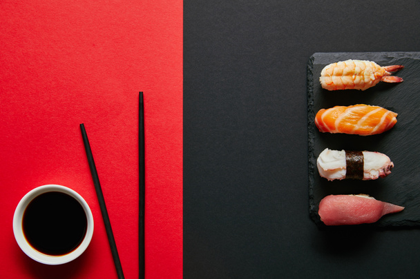 acostado plano con salsa de soja en un tazón, palillos y sushi nigiri establecidos en el plato de pizarra negra sobre fondo rojo y negro
 - Foto, Imagen