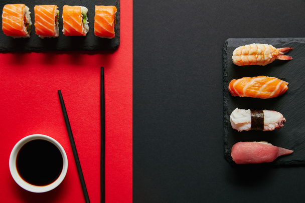 flat lay with soya sauce in bowl, chopsticks and sushi sets on black slate plates on red and black background - Фото, изображение