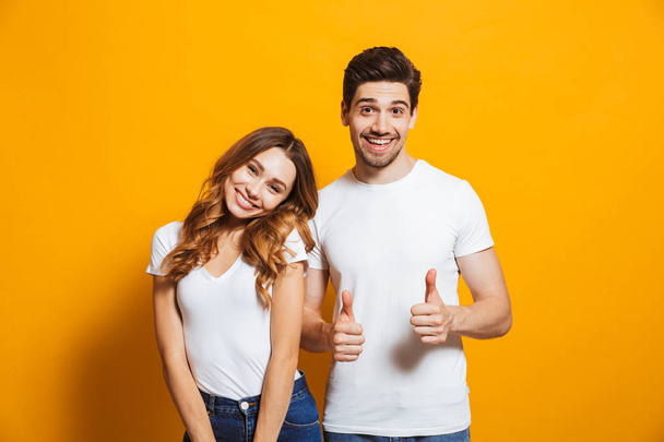 陽気なカップルの男と 20 代の女性基本的な服笑顔と示す親指を黄色背景に分離カメラの肖像画 - 写真・画像