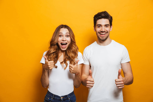 Image of happy young people man and woman in basic clothing laughing and showing thumbs up at camera isolated over yellow background - Zdjęcie, obraz