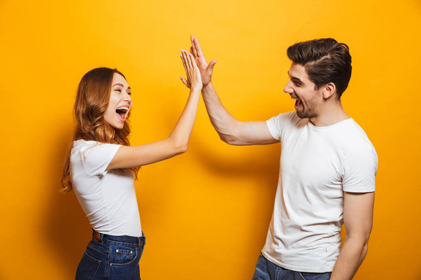 Image of friendly young people man and woman in basic clothing laughing and giving high five isolated over yellow background - Fotó, kép