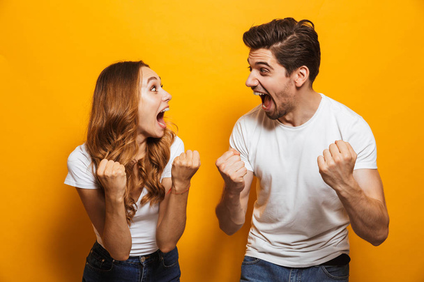 Photo of delighted european man and woman in basic clothing screaming and clenching fists like winners or happy people isolated over yellow background - Foto, imagen