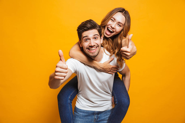 Image of joyous couple showing thumbs up while man piggybacking joyful woman isolated over yellow background - Foto, Imagem