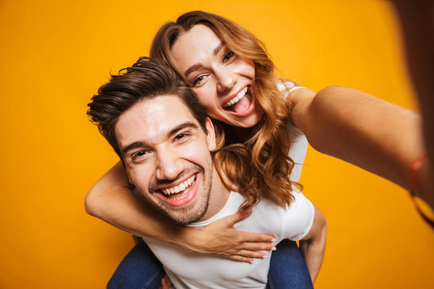 Image of caucasian couple having fun and taking selfie while man piggybacking cute young woman isolated over yellow background - Fotó, kép