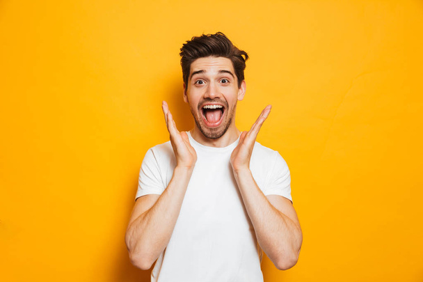 Photo of amazed man in basic clothing screaming in surprise or delight and raising arms isolated over yellow background - Foto, imagen
