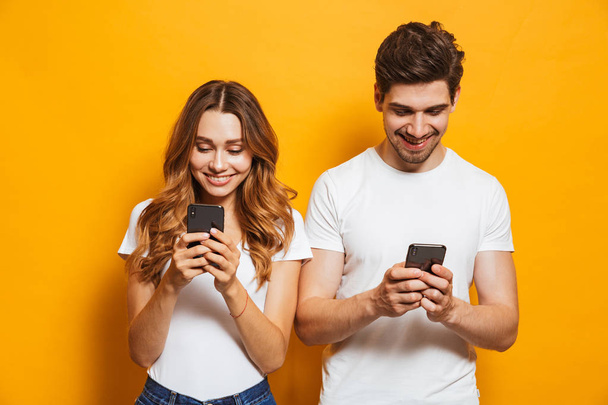 Photo of beautiful people man and woman smiling while both using mobile phones isolated over yellow background - Photo, image