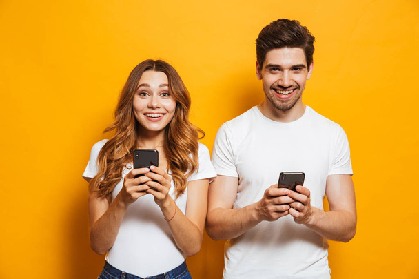 Photo of pleased attractive people man and woman smiling while both using mobile phones, isolated over yellow background - Photo, Image