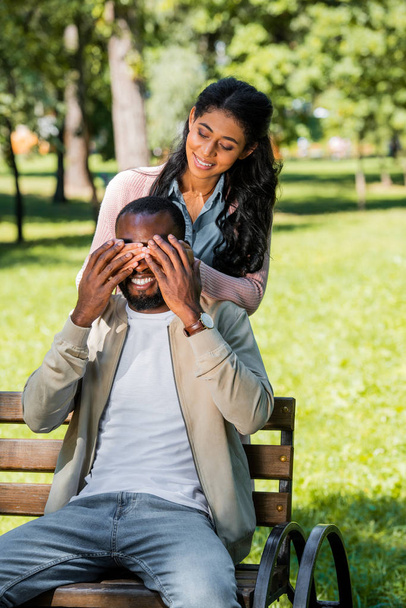 african american girlfriend closing boyfriend eyes while he sitting on bench in park - Фото, зображення