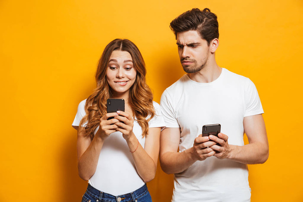 Image of curious man frowning and peeking at smartphone of his girlfriend isolated over yellow background - Φωτογραφία, εικόνα