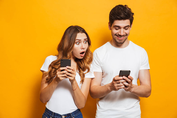 Image of young woman spying and peeking at smartphone of her boyfriend isolated over yellow background - Фото, изображение