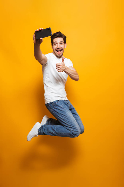 Full length portrait of a joyful young man taking a sefie with mobile phone while jumping isolated over yellow background - Photo, image