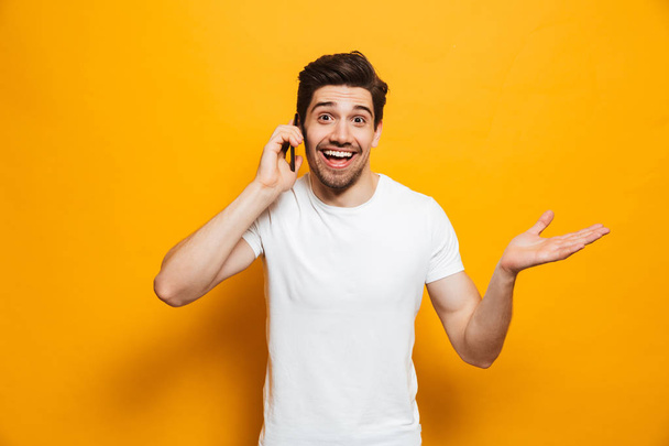 Portrait of a cheerful young man talking on mobile phone isolated over yellow background - 写真・画像